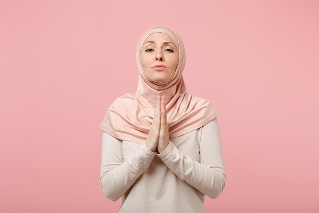 Plädierende junge arabische muslimische Frau in Hijab-leichter Kleidung posiert isoliert auf rosa Hintergrund im Studio. Menschen religiösen Islam Lifestyle-Konzept. Mock-up-Kopienbereich. Händchen haltend zum Gebet gefaltet.