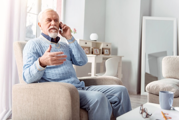 Pläne schmieden. Angenehmer älterer Mann, der im Sessel im Wohnzimmer sitzt und mit seinem Freund telefoniert und über das Abendessen spricht