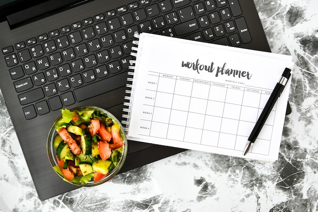 Plan de ejercicios para una semana y tazón con ensalada de verduras en el lugar de trabajo cerca de la computadora.