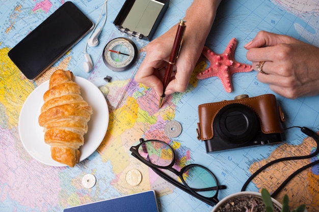 Plan de viaje. antecedentes: qué llevar de viaje. Manos y mapa de mujeres, cámara retro, dinero, monedas, croissant, café, bolígrafo, brújula, gafas de sol, teléfono inteligente