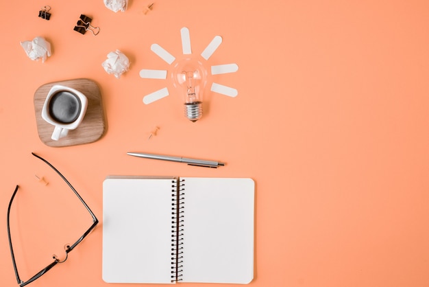 Plana colocar planejamento financeiro brainstorming imagem de mesa desarrumado com placa de grampo em branco, material de escritório, caneta, bloco de notas, óculos, xícara de café, lâmpada em fundo laranja.