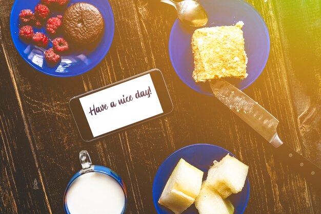 Plana leigos na mesa de madeira com comida no café da manhã com texto no telefone tenha um bom dia. leite, panquecas e frutas a