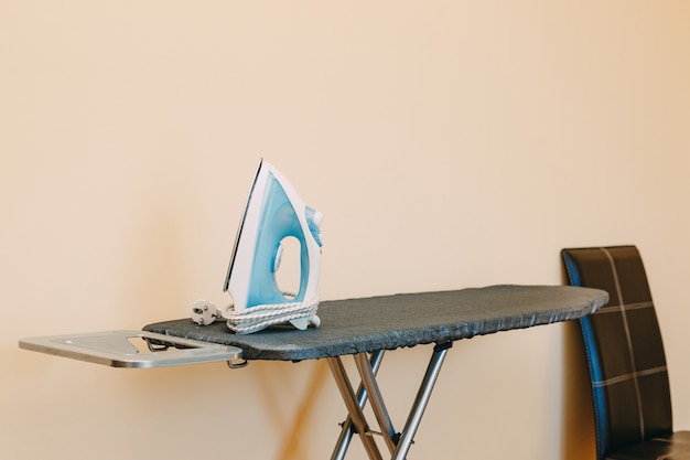 Una plancha azul sobre una tabla de planchar con una funda cerca de la silla en la habitación.