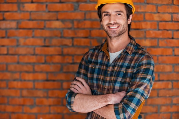 Foto planificación de una nueva construcción. hombre joven sonriente en casco manteniendo los brazos cruzados y mirando a la cámara mientras está de pie contra la pared de ladrillo