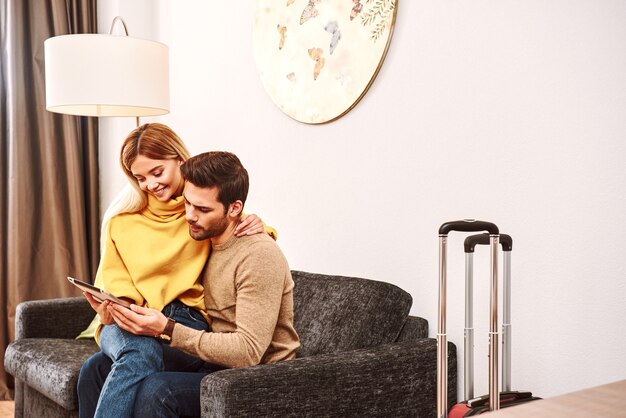 Planificación de vacaciones. Mujer joven sentada en las rodillas de su novio y soñando. Feliz pareja madura con maletas buscando en tableta en la habitación del hotel