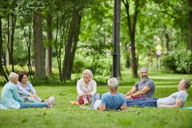 Plano amplio de personas mayores modernas pasar la mañana de verano juntos relajándose en el césped en el parque después de hacer ejercicio