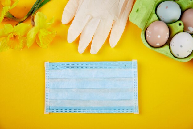 Plano de composición de pascua en la cuarentena de coronavirus. Símbolo de Pascua huevos y flores de narciso cerca de guantes y máscaras médicas protectoras