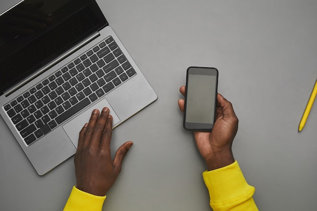 Plano de fundo cinza mínimo de mãos masculinas afro-americanas segurando um smartphone com uma tela em branco sobre o laptop enquanto trabalha na mesa, vista de cima para baixo, copie o espaço