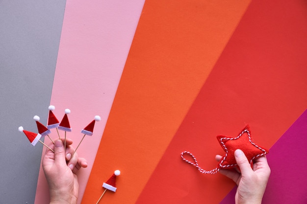 Plano de natal leigos com mãos femininas segurando estrela têxtil e pequenos chapéus de papai noel. vista superior do fundo de papel em camadas geométrico multicolor nas camadas vermelhas, laranja, rosa, magenta e prata de cima.