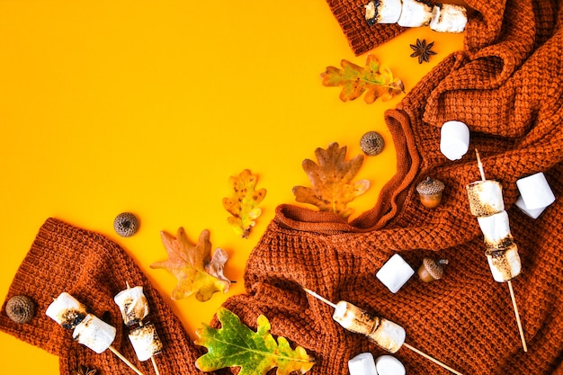 Plano de outono deitado com suéter laranja de malha, marshmallows grelhados e folhas em fundo amarelo, vista superior. Copie o espaço para o texto. Ornamento de Ação de Graças. Quadro de outono