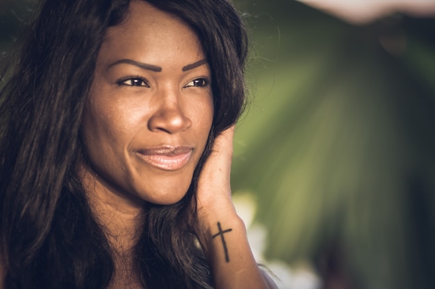 Plano detallado del rostro de una hermosa joven caribeña - Fondo colorido y borroso - Tatuaje de cruz en la mano - Primer plano