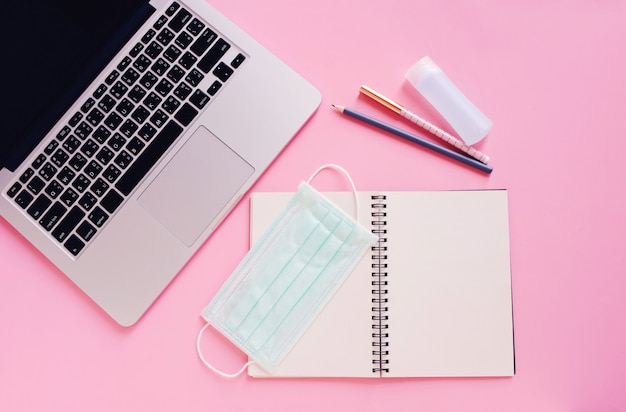 Plano del escritorio del espacio de trabajo con computadora portátil, cuaderno en blanco, máscaras médicas y gel de alcohol en rosa brillante