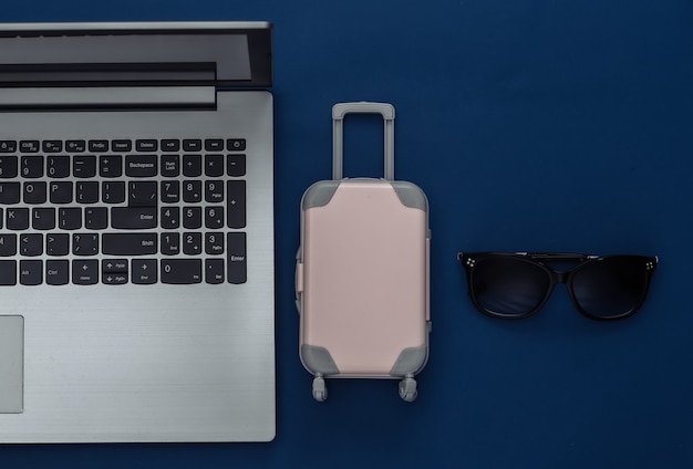 Plano leigo férias férias e conceito de planejamento de viagens. Laptop e mini mala de viagem de plástico, passaporte, óculos de sol sobre fundo azul clássico. Vista do topo