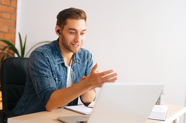 Plano medio de un apuesto joven hombre de negocios con un auricular que tiene una videoconferencia en línea en una laptop en la oficina de casa. Un estudiante confiado ve un curso de formación de seminarios web en una computadora sentada en el escritorio.