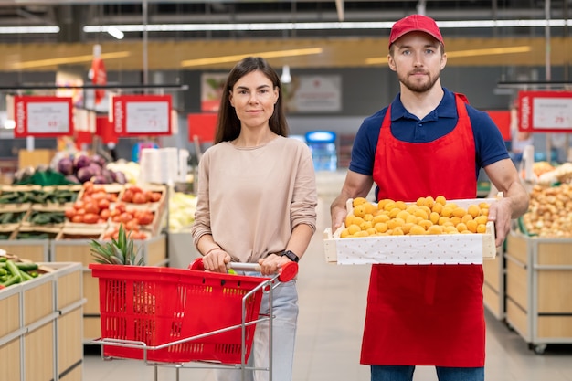 Plano medio horizontal de trabajador varón sosteniendo una caja de frutas de pie con una joven clienta en el supermercado