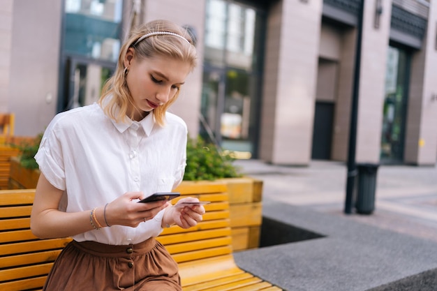 Plano medio de una joven enfocada ingresando el número de tarjeta de crédito para comprar en línea usando un teléfono inteligente sentado en un banco amarillo en la calle de la ciudad en el día de verano