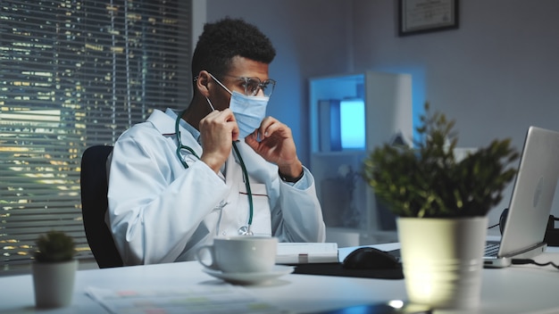 Plano medio del joven médico africano que muestra cómo usar una máscara médica por video llamada en la computadora