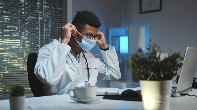 Plano medio del joven médico africano que muestra cómo usar una máscara médica por video llamada en la computadora