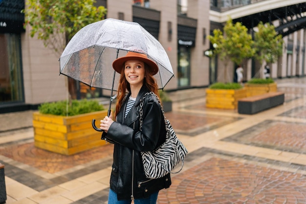 Plano medio de una joven pelirroja positiva con sombrero de moda caminando por la calle de la ciudad europea con un paraguas transparente disfrutando del clima lluvioso al aire libre