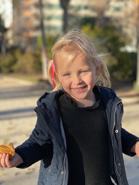 Plano medio de una niña pequeña alegre con un bollo en la mano. Retrato al aire libre de un niño de ojos azules con cabello rubio.