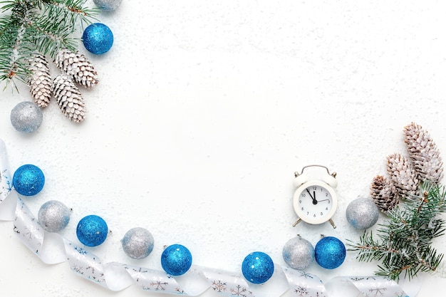 Plano de Navidad con reloj despertador blanco, ramas de nieve de abeto azul, conos de abeto, bolas de Navidad azules y plateadas y cinta blanca sobre blanco