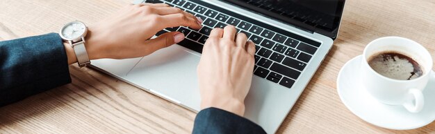 Foto plano panorámico de empresaria escribiendo en la computadora portátil cerca de la taza con café en la mesa