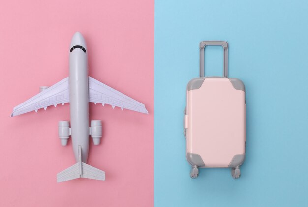 Plano de viaje aéreo. Mini maleta de viaje de plástico y avión sobre fondo rosa pastel azul. Vista superior.