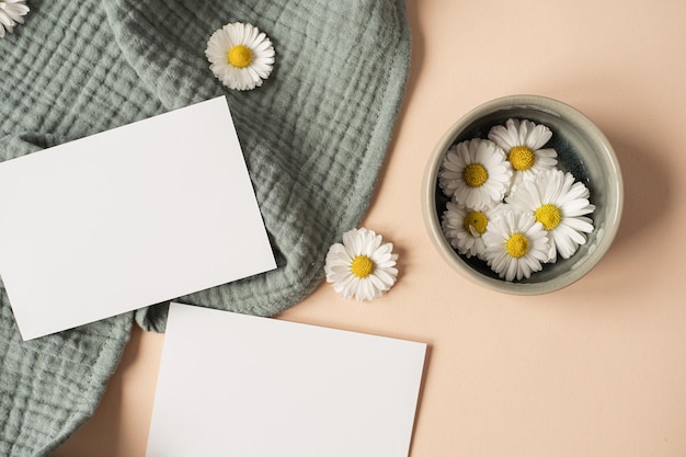 Plantilla de invitación de estilo parisino estético Flatlay Tarjeta de hoja de papel en blanco con capullos de manzanilla de espacio de copia de maqueta y tela de muselina aguamarina sobre fondo beige neutro Vista superior plana