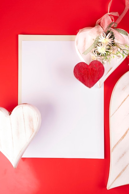 Plantilla de tarjeta de San Valentín con hoja de papel en blanco y dos corazones de madera pintados con arreglos florales hechos a mano sobre fondo rojo.