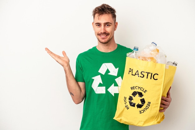 Plástico reciclado jovem homem caucasiano isolado no fundo branco, mostrando um espaço de cópia na palma da mão e segurando a outra mão na cintura.