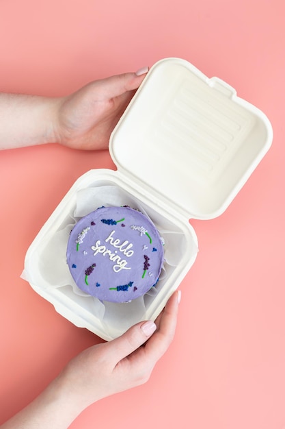 Plastik-Lunchbox mit köstlichem Bento-Kuchen Hallo Frühling in Frauenhänden auf rosa Hintergrund, Draufsicht