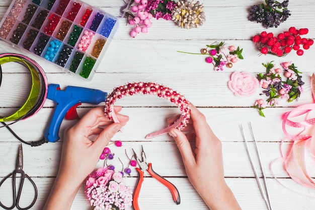 Plastikbeeren, Blumen, Perlen und Instrumente zum Anfertigen von handgefertigten Stirnbändern. Draufsicht mit den Händen