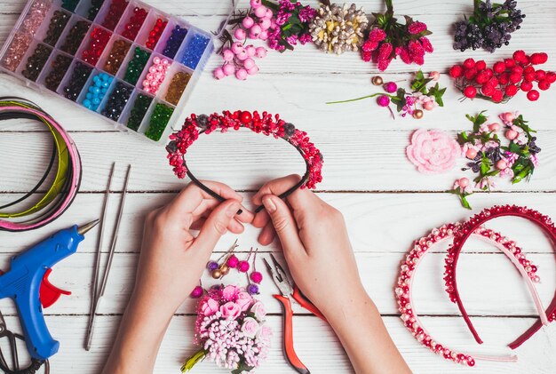 Plastikbeeren, Blumen, Perlen und Instrumente zum Anfertigen von handgefertigten Stirnbändern. Draufsicht mit den Händen