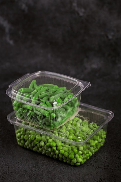 Plastikbehälter mit verschiedenen Bio-Tiefkühlgemüse auf schwarzem Tisch. Grüne Erbsen und geschnittene grüne Bohnen in einer Box