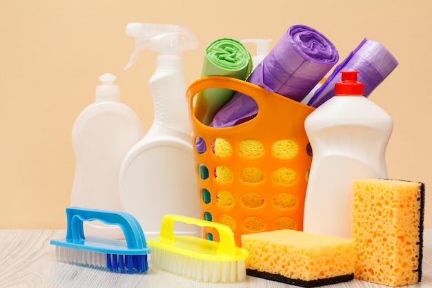 Plastikflaschen mit Glas- und Fliesenreiniger, orangefarbener Korb mit Müllsäcken, Schwämmen, Bürsten auf beigem Hintergrund. Wasch- und Reinigungsset.