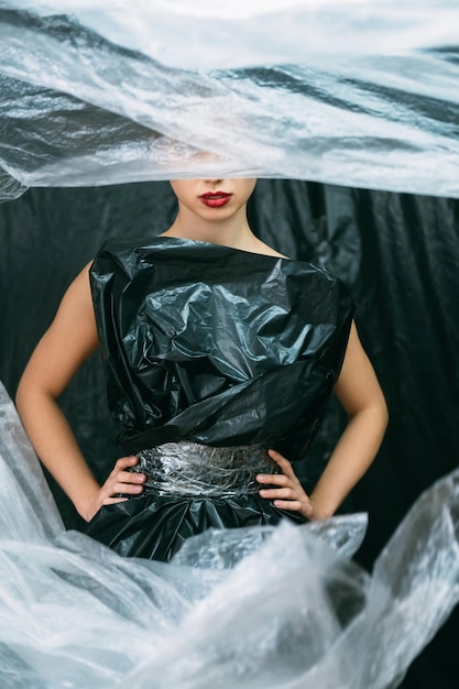 Plastikmode Abfallreduzierung Naturschutz Luftverschmutzung Weibliches Modell mit roten Lippen, das in schwarzem Müllsackkleid posiert, isoliert mit weißer Polyethylenfolie auf dunklem, zerknittertem Hintergrund