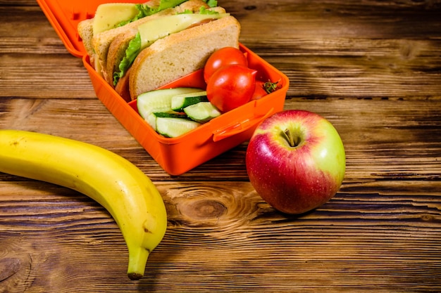 Plátano de manzana madura y caja de almuerzo con sándwiches de pepinos y tomates en la mesa de madera