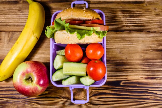 Plátano de manzana maduro y caja de almuerzo con pepinos de hamburguesa y tomates en la mesa de madera rústica Vista superior