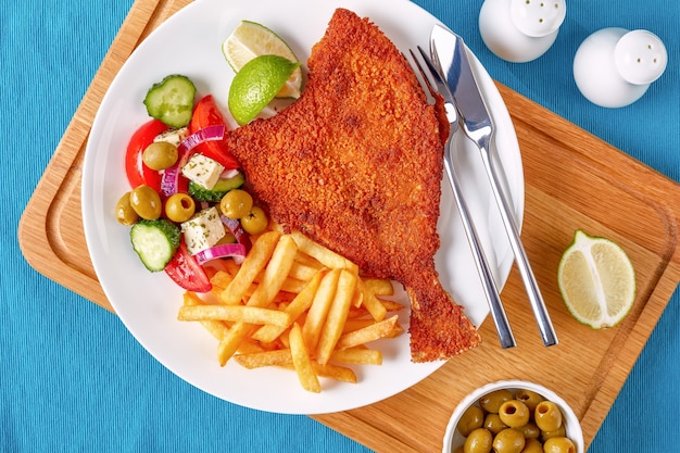 Platija frita crujiente en pan rallado servida con verduras frescas, queso feta, aceitunas, ensalada griega y papas fritas en un plato blanco con tenedor y cuchillo de plata sobre una tabla de cortar, vista desde arriba