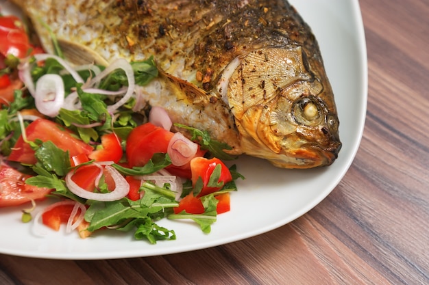 Platillo de pescado. Carpa cruciana asada en un plato blanco con vegetales frescos de la mezcla en la tabla. El primer frió el carassius listo para comer. Plato caliente Plato principal.