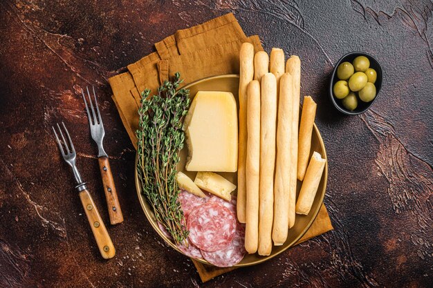 Plato de aperitivo con aperitivos antipasti grissini palitos de pan salami aceitunas y queso parmesano Bakground oscuro Vista superior