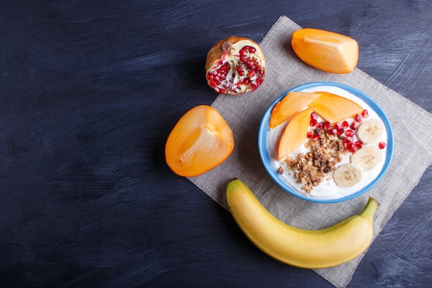 Un plato azul con yogur griego, granola, caqui, plátano, granada.