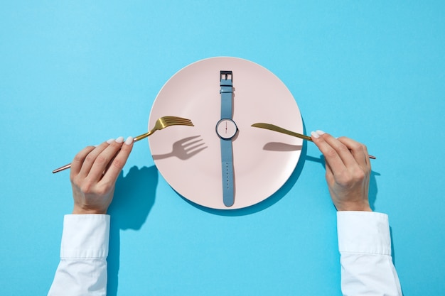 El plato blanco con banda azul muestra a las seis en punto servido cuchillo y tenedor en las manos de una niña en una pared azul, lugar para el texto. Hora de comer y concepto de dieta. Vista superior.