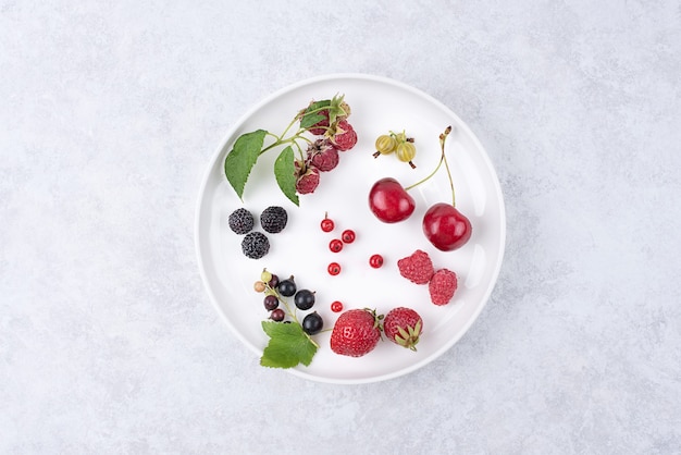 Plato blanco con bayas, fresas, frambuesas, grosellas, cerezas sobre un fondo blanco, vista superior.