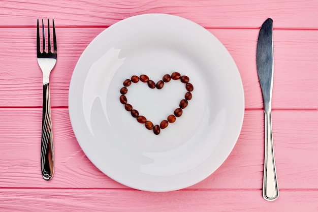 Foto plato blanco con corazón de granos de café. placa blanca con granos de café en forma de corazón, tenedor y cuchillo sobre fondo de madera rosa, vista superior.