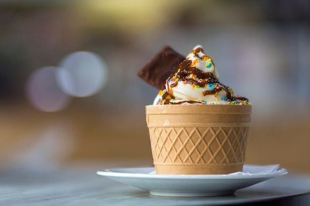 Plato blanco con postre de helado en taza de oblea con galletas de chocolate y decoración creativa relleno en fondo interior colorido borroso.