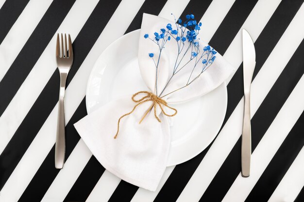 Un plato blanco vacío con una servilleta y flores en forma de arco y un cuchillo tenedor sobre la mesa de rayas blancas negras