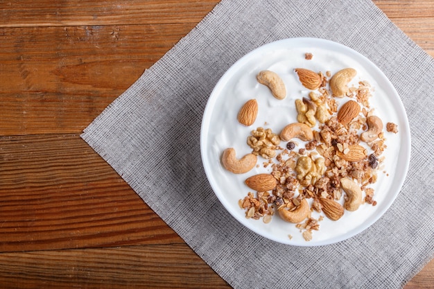 Plato blanco con yogur griego granola almendra anacardos nueces sobre fondo de madera marrón