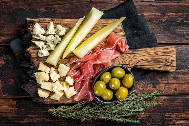 Plato de carne y queso antipasti con jamón Prosciutto, parmesano, queso azul, melón y aceitunas sobre tabla de madera, fondo de madera, vista superior