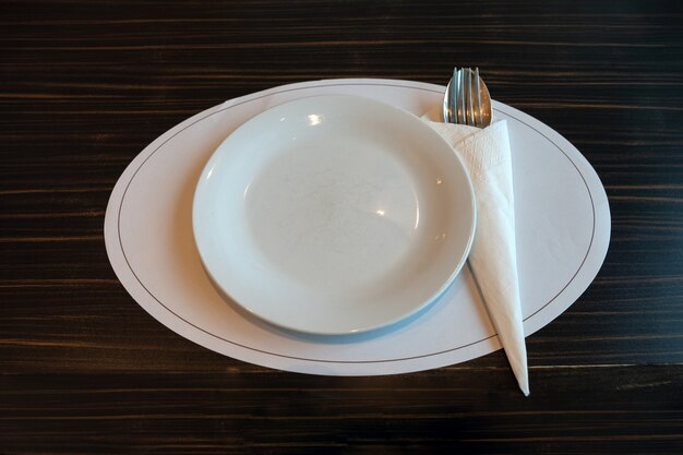 Plato de cerámica blanca con cuchara, tenedor y servilleta en la mesa de madera en el restaurante